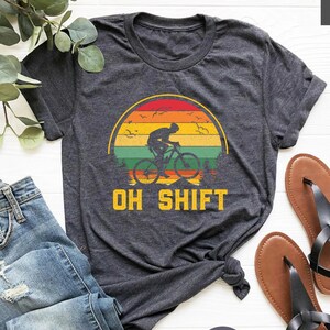 May include: Dark gray t-shirt with a colorful sunset design featuring a cyclist and the text "OH SHIFT". The shirt is paired with blue jeans and brown sandals. The shirt is made of a soft, comfortable material.