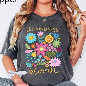 May include: A charcoal gray short-sleeve t-shirt featuring a colorful floral graphic print. The text reads Let Kindness Bloom in yellow script. The design includes various stylized flowers in shades of blue, pink, yellow, and green.