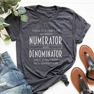 May include: Dark gray t-shirt with the text "THERE IS A FINE LINE BETWEEN NUMERATOR AND DENOMINATOR ONLY A FRACTION WILL UNDERSTAND." The shirt is paired with blue jeans and brown sandals with black straps. A sprig of greenery is in the background.