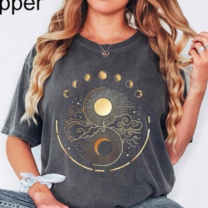 May include: Pepper-colored t-shirt featuring a gold celestial design with a sun, moon, and phases of the moon. The graphic is centered on the front of the shirt. The shirt has short sleeves and a relaxed fit.