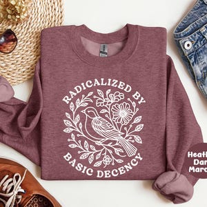 May include: A heather dark maroon crewneck sweatshirt featuring a white graphic print of a bird perched on a branch surrounded by flowers and leaves, with the text "RADICALIZED BY BASIC DECENCY" arched around the central illustration.