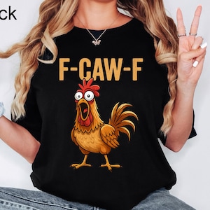 May include: Black t-shirt with the text "F-CAW-F" in gold letters above a cartoon rooster illustration. The rooster is brown, orange, and red with an open beak. The word "Black" is in the upper left corner.