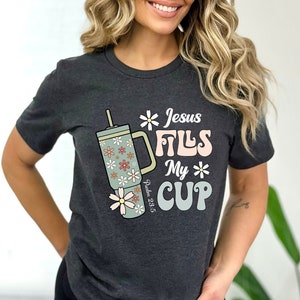 May include: A dark gray t-shirt with a graphic of a floral patterned tumbler with the text "Jesus Fills My Cup" and "Psalm 23:5" printed on it.