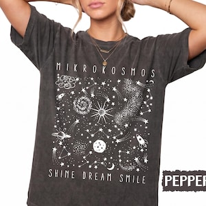 May include: A charcoal gray short-sleeve t-shirt featuring a white graphic print of space elements including stars, planets, rockets, and a galaxy. The text MIKROKOSMOS is printed above the graphic, and SHINE DREAM SMILE is printed below it.