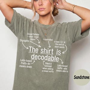 May include: A sandstone-colored short-sleeve t-shirt featuring white text that reads The shirt is decodable, surrounded by arrows pointing to linguistic explanations including Closed Syllable, Digraphs, r-controlled, and Latin-based suffixes.