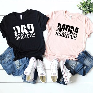 May include: Two black t-shirts with white text and dinosaur silhouettes. The text on the black shirt says "DAD asaurus". The text on the pink shirt says "MOM asaurus".