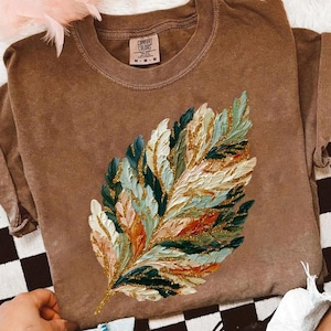 May include: A brown Comfort Colors t-shirt featuring a large, colorful leaf design. The leaf is painted with textured brushstrokes in shades of green, blue, orange, and gold. The shirt has short sleeves and a crew neck.