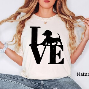 May include: Cream-colored t-shirt with the word "LOVE" in large black letters. A black silhouette of a dachshund dog is positioned above the "VE". The word "Natural" is printed in black on the lower right.