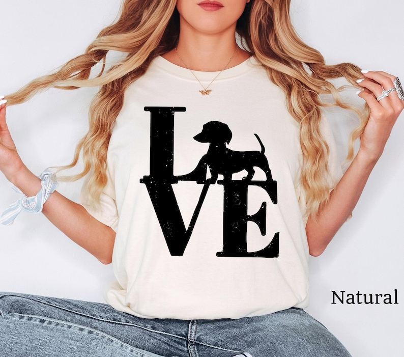 May include: Cream-colored t-shirt with the word "LOVE" in large black letters. A black silhouette of a dachshund dog is positioned above the "VE". The word "Natural" is printed in black on the lower right.