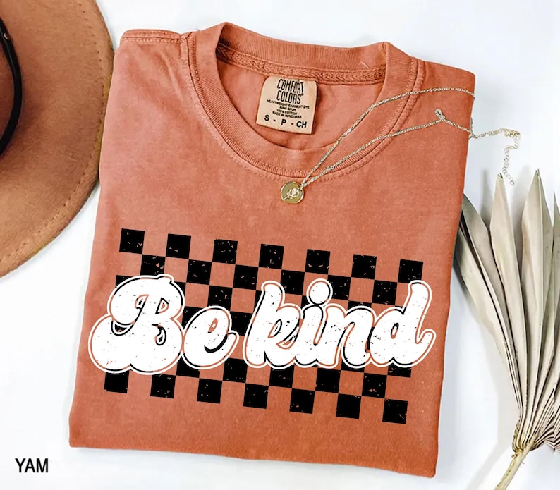 Pu&ograve; includere: Una t-shirt a maniche corte color terracotta con una grafica a scacchi nera e terracotta e la scritta bianca "Be kind" in un carattere grassetto e arrotondato. La maglietta presenta un girocollo e un'etichetta del marchio Comfort Colors.