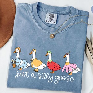 May include: Blue jean Comfort Colors t-shirt with a whimsical illustration of four geese dressed in various outfits. The phrase "just a silly goose" is printed in white script below the geese. A gold necklace is draped across the shirt.