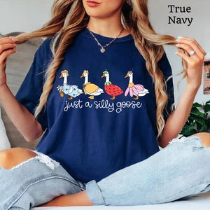 May include: Navy blue t-shirt with a whimsical design featuring four geese dressed in various outfits, including a floral dress, a cape, and a strawberry costume. The text "just a silly goose" is printed below the geese.