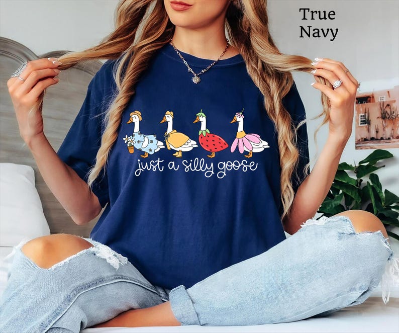 May include: Navy blue t-shirt with a whimsical design featuring four geese dressed in various outfits, including a floral dress, a cape, and a strawberry costume. The text "just a silly goose" is printed below the geese.