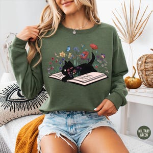 May include: Olive green sweatshirt featuring a black cat lying on an open book, surrounded by colorful flowers and butterflies. The graphic design includes red poppies, yellow daffodils, and blue butterflies. The sweatshirt has long sleeves and a crew neck.