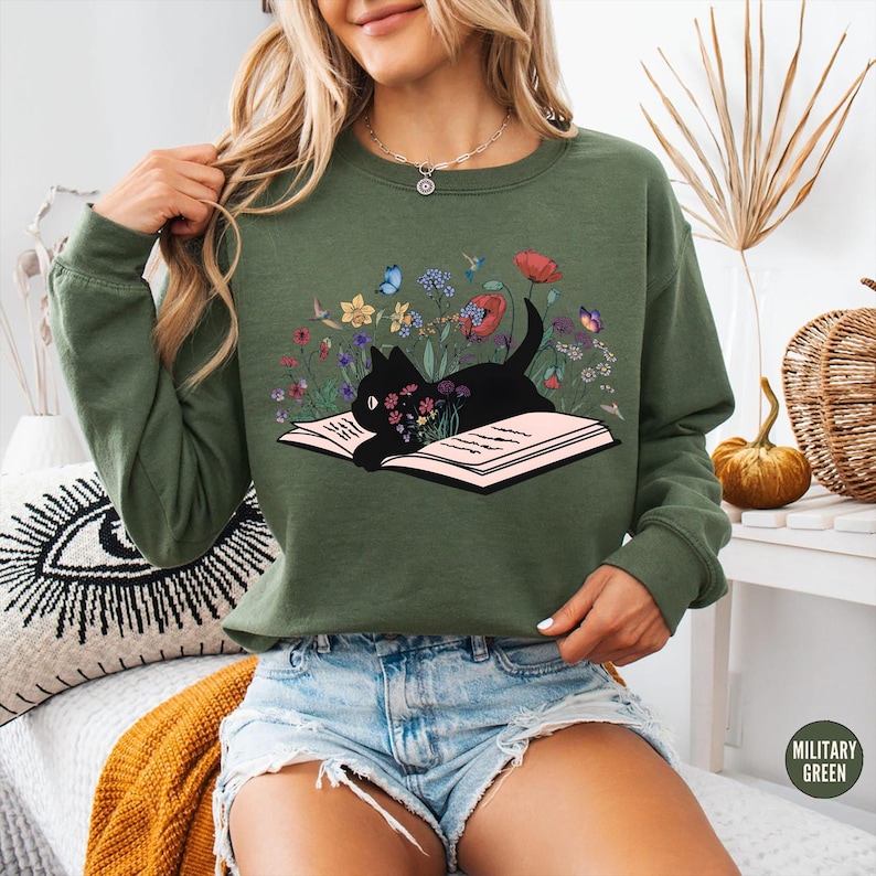 May include: Olive green sweatshirt featuring a black cat lying on an open book, surrounded by colorful flowers and butterflies. The graphic design includes red poppies, yellow daffodils, and blue butterflies. The sweatshirt has long sleeves and a crew neck.
