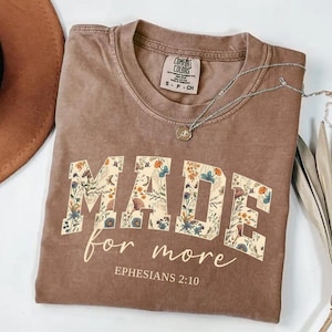 May include: A brown Comfort Colors t-shirt with the words "MADE for more" in a floral design. The text "Ephesians 2:10" is below the main text. A silver necklace with a pendant is on the shirt.