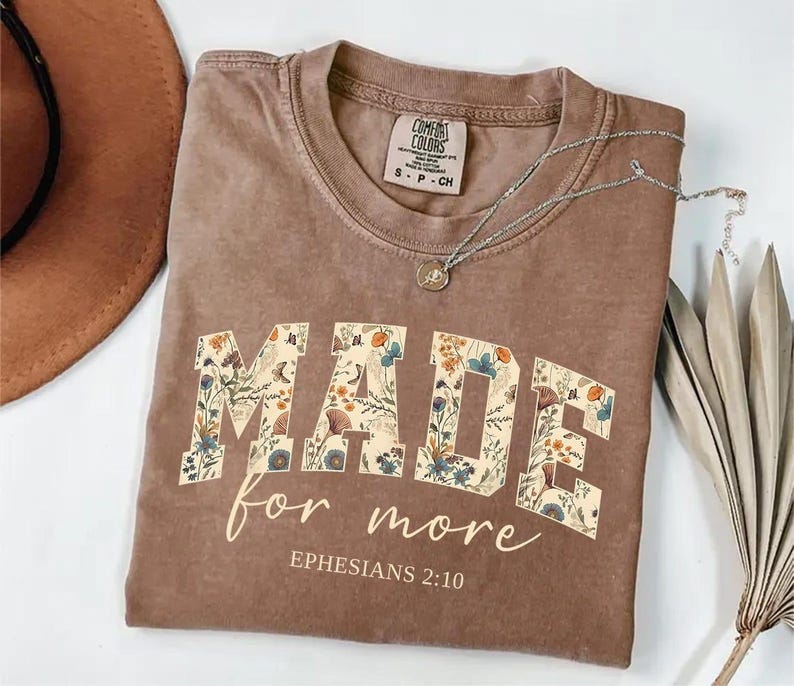 May include: A brown Comfort Colors t-shirt with the words "MADE for more" in a floral design. The text "Ephesians 2:10" is below the main text. A silver necklace with a pendant is on the shirt.