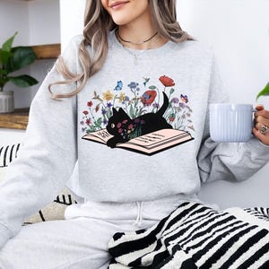 May include: A light gray sweatshirt featuring a black cat lying on an open book, surrounded by colorful flowers and butterflies. The cat's body is filled with flowers. The sweatshirt is part of a matching set with gray sweatpants.