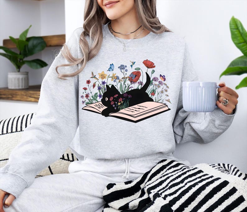 May include: A light gray sweatshirt featuring a black cat lying on an open book, surrounded by colorful flowers and butterflies. The cat's body is filled with flowers. The sweatshirt is part of a matching set with gray sweatpants.
