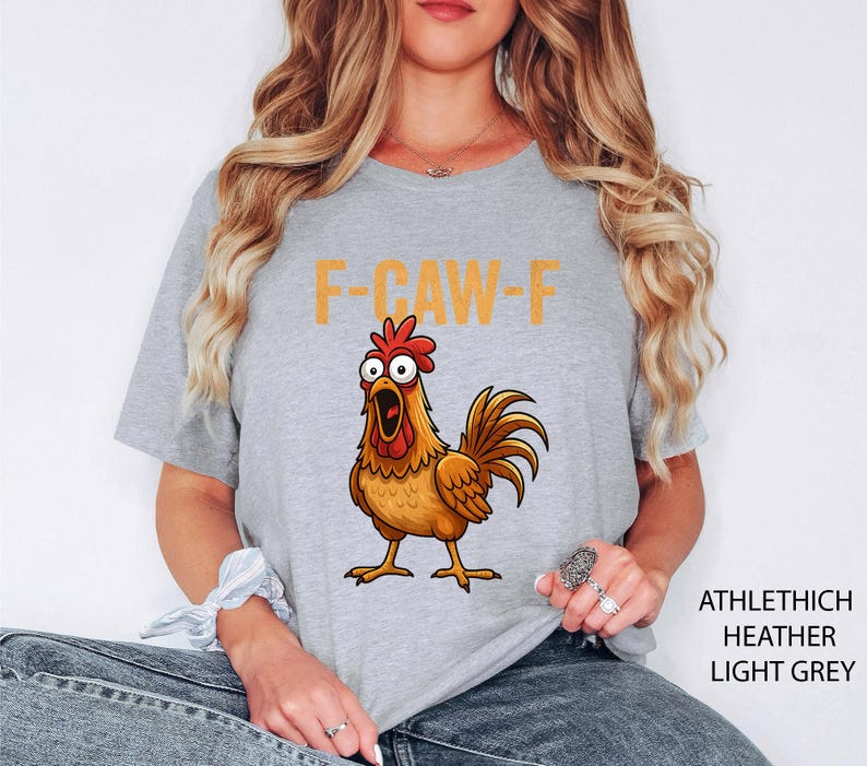 May include: Light grey t-shirt with a cartoon chicken graphic and the orange text "F-CAW-F". The shirt is made of a soft, heathered material. The text "ATHLETHICH HEATHER LIGHT GREY" is printed on the right side.