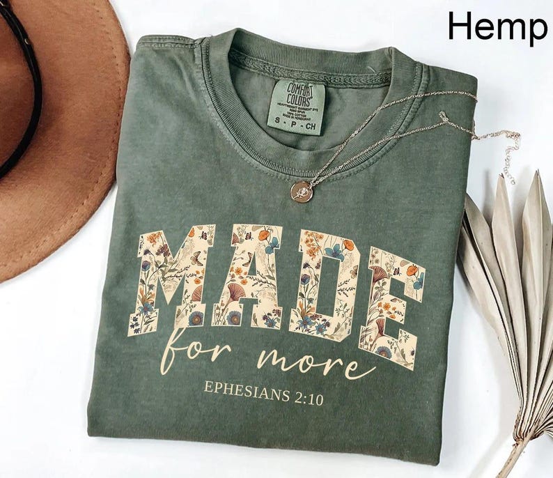 May include: A green Comfort Colors t-shirt with the words "MADE for more" in a floral design. The text "Ephesians 2:10" is below the main text. A silver necklace with a small pendant is on the shirt.