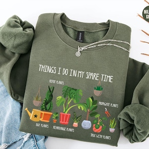 May include: Military green sweatshirt with the text "THINGS I DO IN MY SPARE TIME" and illustrations of plants. The design includes a watering can, potted plants, and the phrases "REPOT PLANTS", "BUY PLANTS", "REARRANGE PLANTS", "PROPAGATE PLANTS", and "TALK WITH PLANTS".