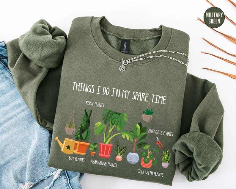 May include: Military green sweatshirt with the text "THINGS I DO IN MY SPARE TIME" and illustrations of plants. The design includes a watering can, potted plants, and the phrases "REPOT PLANTS", "BUY PLANTS", "REARRANGE PLANTS", "PROPAGATE PLANTS", and "TALK WITH PLANTS".