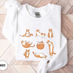 May include: A white crewneck sweatshirt featuring a graphic print of seven orange and white tabby cats in various poses, including sitting, stretching, and curled up. The garment has a ribbed collar and long sleeves with a Gildan brand label inside.