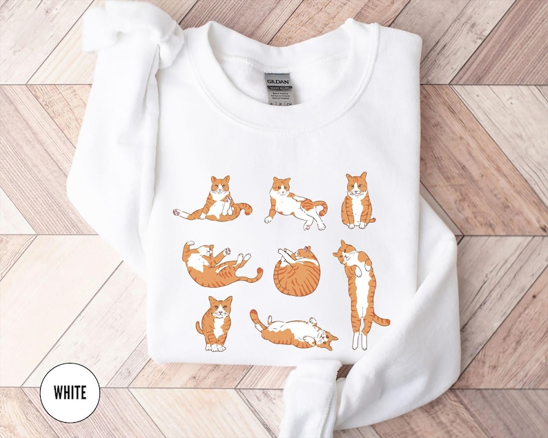 May include: A white crewneck sweatshirt featuring a graphic print of seven orange and white tabby cats in various poses, including sitting, stretching, and curled up. The garment has a ribbed collar and long sleeves with a Gildan brand label inside.