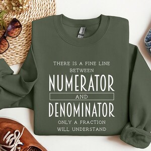 May include: An olive green sweatshirt with white text that reads "THERE IS A FINE LINE BETWEEN NUMERATOR AND DENOMINATOR ONLY A FRACTION WILL UNDERSTAND." The sweatshirt is displayed on a white surface with brown shoes, sunglasses, and denim jeans.