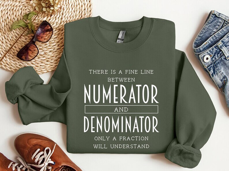 May include: An olive green sweatshirt with white text that reads "THERE IS A FINE LINE BETWEEN NUMERATOR AND DENOMINATOR ONLY A FRACTION WILL UNDERSTAND." The sweatshirt is displayed on a white surface with brown shoes, sunglasses, and denim jeans.
