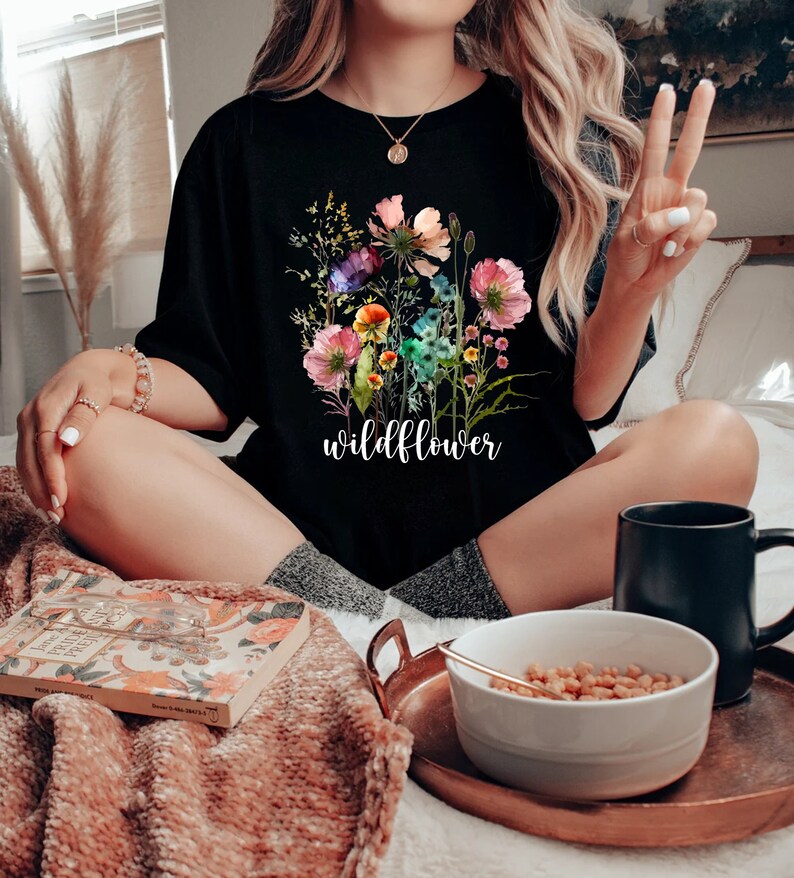 May include: Black t-shirt featuring a colorful wildflower bouquet design with the word "wildflower" in white script. The floral arrangement includes pink, purple, and yellow flowers. The shirt is a casual style.