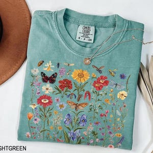May include: Light green t-shirt with a floral print featuring butterflies, bees, and other insects. The shirt has a crew neck and short sleeves. The text "COMFORT COLORS" is printed on the tag.