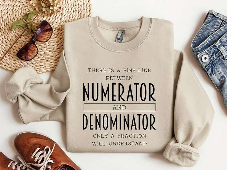 May include: A beige sweatshirt with the text "THERE IS A FINE LINE BETWEEN NUMERATOR AND DENOMINATOR ONLY A FRACTION WILL UNDERSTAND" in black. The sweatshirt is displayed on a white surface with sunglasses, shoes, and jeans.