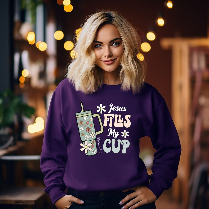 K&ouml;nnte beinhalten: Ein lila Sweatshirt mit dem Text "Jesus Fills My Cup" und einer floralen Bechergrafik. Der Becher ist hellgr&uuml;n mit wei&szlig;en und orangefarbenen Blumen. Das Sweatshirt enth&auml;lt auch den Bibelvers Psalm 23:5.