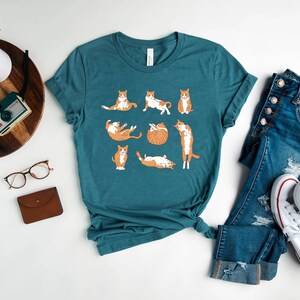 May include: Teal t-shirt featuring a playful design of orange and white cats in various poses. The shirt is paired with blue jeans and white sneakers. A wooden tray with a plant, camera, glasses, and a small brown wallet is also visible.