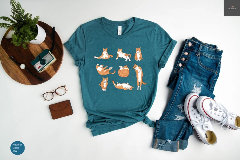 May include: Teal t-shirt featuring a playful design of orange and white cats in various poses. The shirt is paired with blue jeans and white sneakers. A wooden tray with a plant, camera, glasses, and a small brown wallet is also visible.