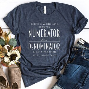 May include: Dark blue t-shirt with white text: "THERE IS A FINE LINE BETWEEN NUMERATOR AND DENOMINATOR ONLY A FRACTION WILL UNDERSTAND." The shirt is styled with blue jeans and beige open-toe shoes. The shirt is made of a soft material.