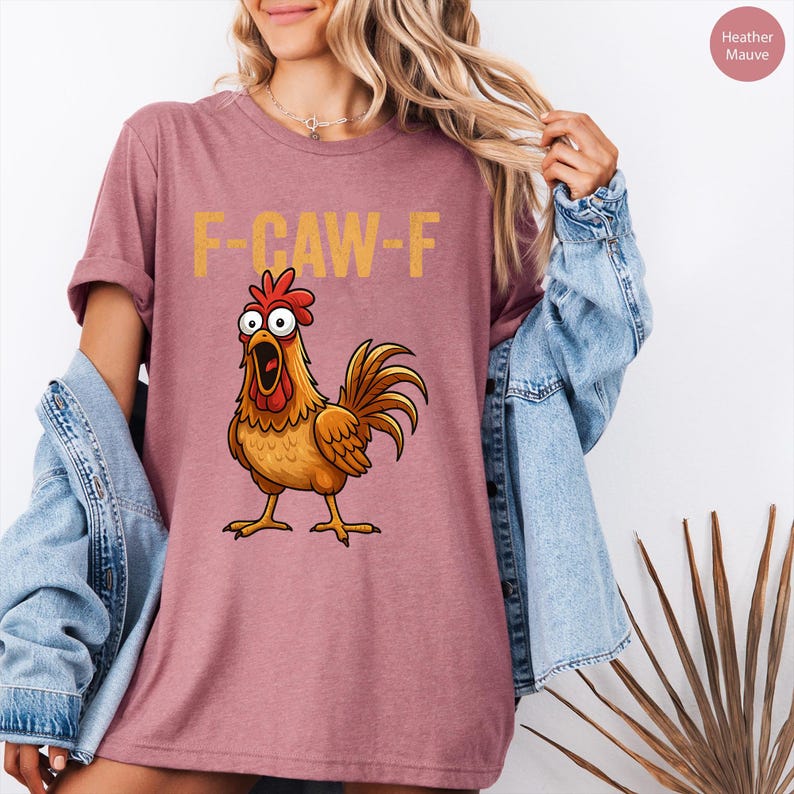 May include: Mauve t-shirt with the text "F-CAW-F" in yellow. The shirt features a cartoon rooster with wide eyes and an open beak. The rooster is brown, orange, and red.