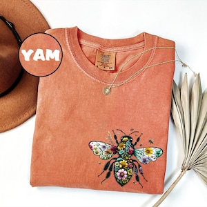 May include: Coral-colored Comfort Colors t-shirt featuring a detailed floral bee graphic. The shirt has a crew neck and short sleeves. A gold necklace with a pendant is draped across the shirt. A brown hat and dried palm frond are also in the image.