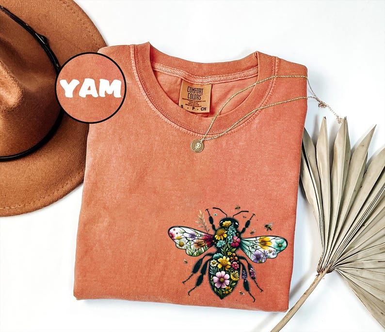 May include: Coral-colored Comfort Colors t-shirt featuring a detailed floral bee graphic. The shirt has a crew neck and short sleeves. A gold necklace with a pendant is draped across the shirt. A brown hat and dried palm frond are also in the image.