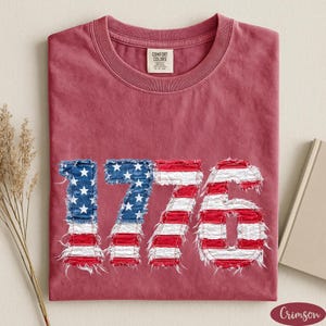 May include: Crimson-colored t-shirt with the number "1776" in a distressed, patriotic design. The numbers are made of denim with white stars and red and white stripes, evoking the American flag.