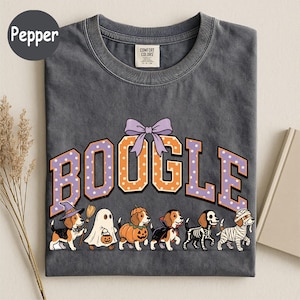 K&ouml;nnte beinhalten: Ein anthrazitfarbenes T-Shirt mit dem Schriftzug BOOGLE in orangefarbenen und violetten gepunkteten Buchstaben und einer violetten Schleife dar&uuml;ber. Darunter sind sechs Beagle-Hunde in Halloween-Kost&uuml;men abgebildet: Hexe, Geist, K&uuml;rbis, Vampir, Skelett und Mumie.