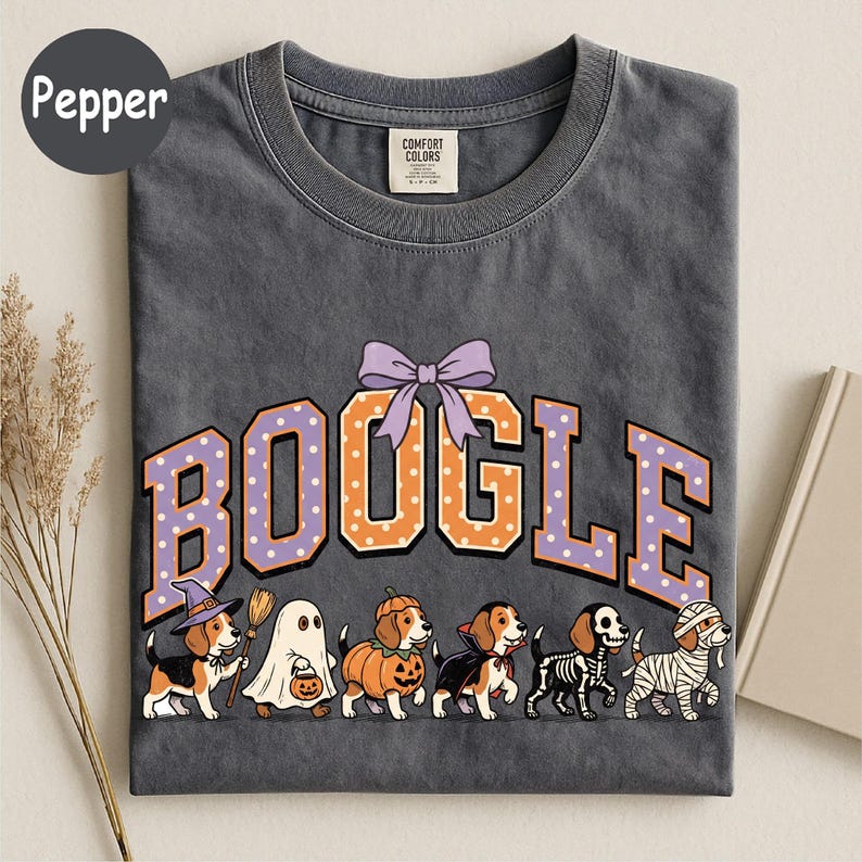 K&ouml;nnte beinhalten: Ein anthrazitfarbenes T-Shirt mit dem Schriftzug BOOGLE in orangefarbenen und violetten gepunkteten Buchstaben und einer violetten Schleife dar&uuml;ber. Darunter sind sechs Beagle-Hunde in Halloween-Kost&uuml;men abgebildet: Hexe, Geist, K&uuml;rbis, Vampir, Skelett und Mumie.