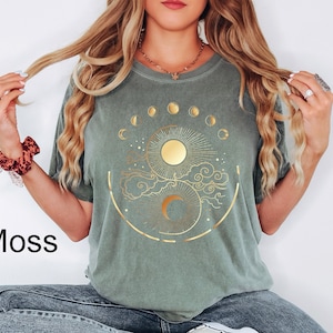 Pu&ograve; includere: Una maglietta verde muschio con un design celeste dorato raffigurante un sole, le fasi lunari e nuvole. La parola "Moss" &egrave; stampata in nero in basso a sinistra. La maglietta ha maniche corte e girocollo.