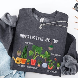 May include: Dark heather gray sweatshirt with the text "THINGS I DO IN MY SPARE TIME" and illustrations of plants. The sweatshirt also features the phrases "REPOT PLANTS", "PROPAGATE PLANTS", "BUY PLANTS", "REARRANGE PLANTS", and "TALK WITH PLANTS".