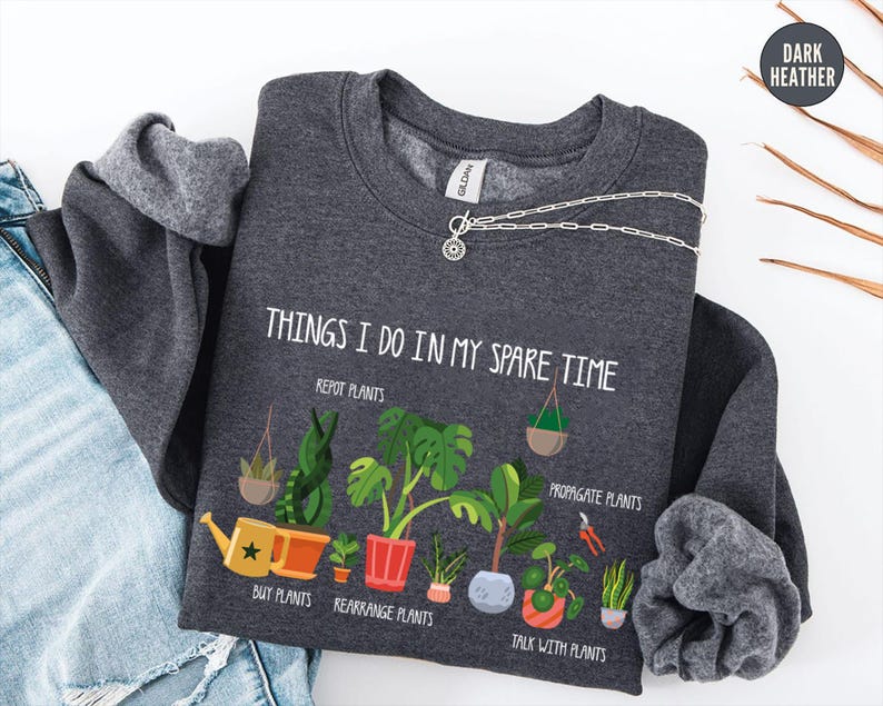 May include: Dark heather gray sweatshirt with the text "THINGS I DO IN MY SPARE TIME" and illustrations of plants. The sweatshirt also features the phrases "REPOT PLANTS", "PROPAGATE PLANTS", "BUY PLANTS", "REARRANGE PLANTS", and "TALK WITH PLANTS".