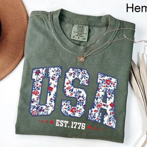 May include: Olive green t-shirt with the word "USA" in a floral pattern, outlined in blue. The text "EST. 1776" is below the word "USA". A silver necklace with a pendant is on the shirt.