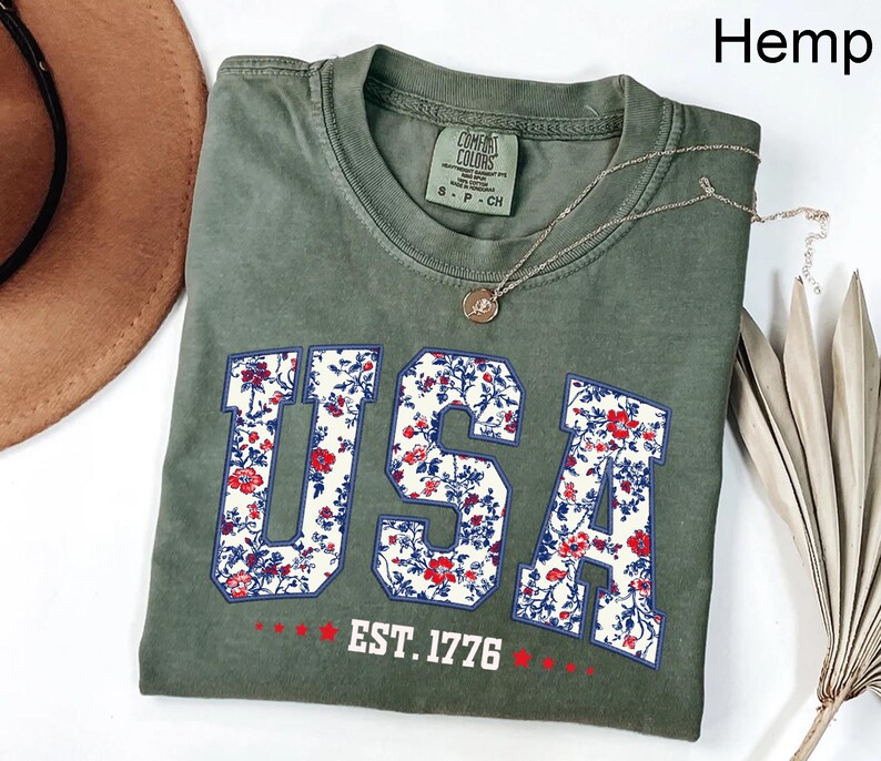 May include: Olive green t-shirt with the word "USA" in a floral pattern, outlined in blue. The text "EST. 1776" is below the word "USA". A silver necklace with a pendant is on the shirt.