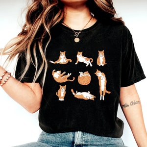 May include: Black t-shirt featuring a pattern of orange and white cats in various poses. The cats are depicted sitting, stretching, and curled up. The shirt is a comfortable, casual style.